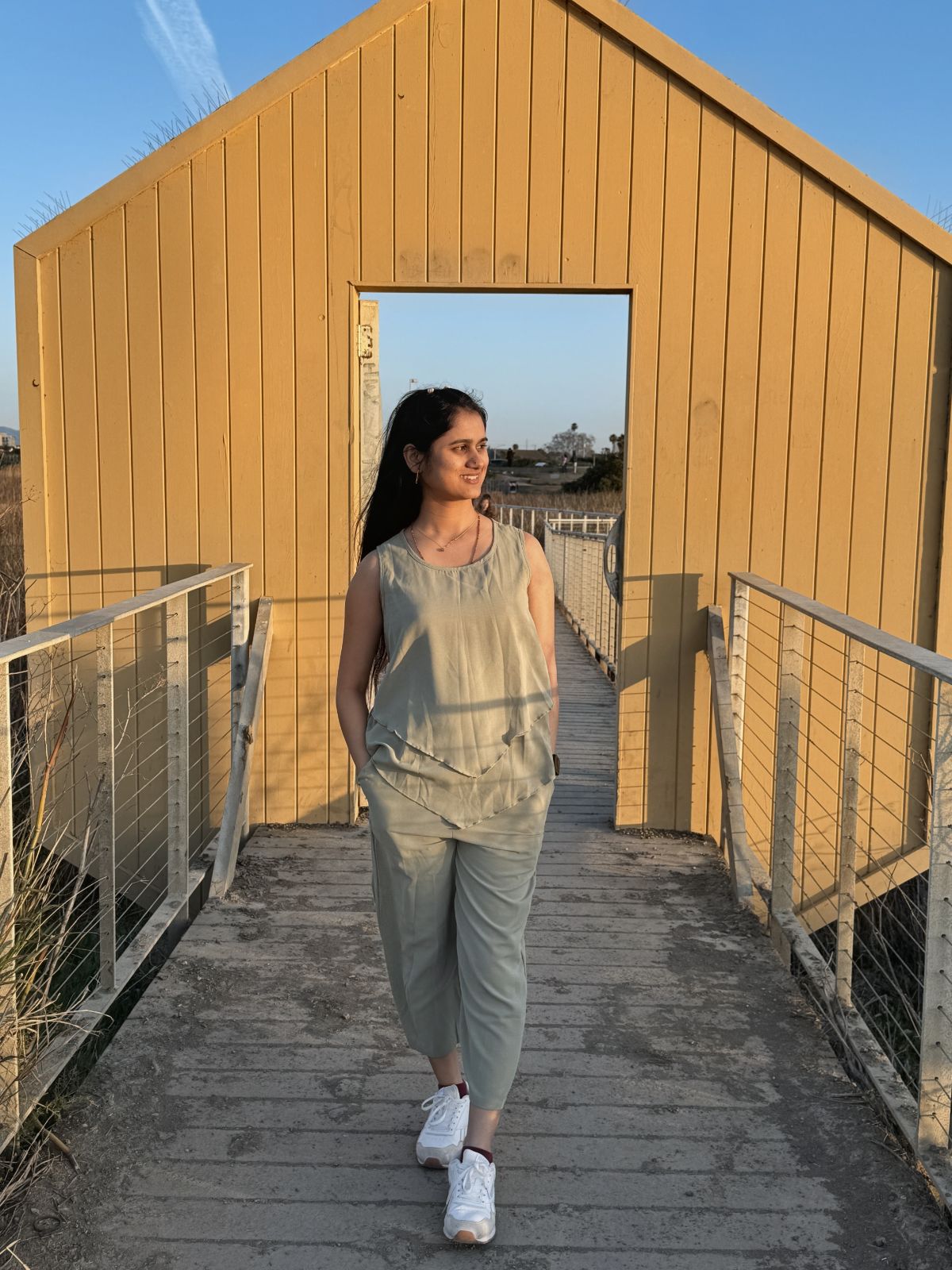 Prajakta Pohare standing on a boardwalk in front of a yellow wooden building at golden hour.
